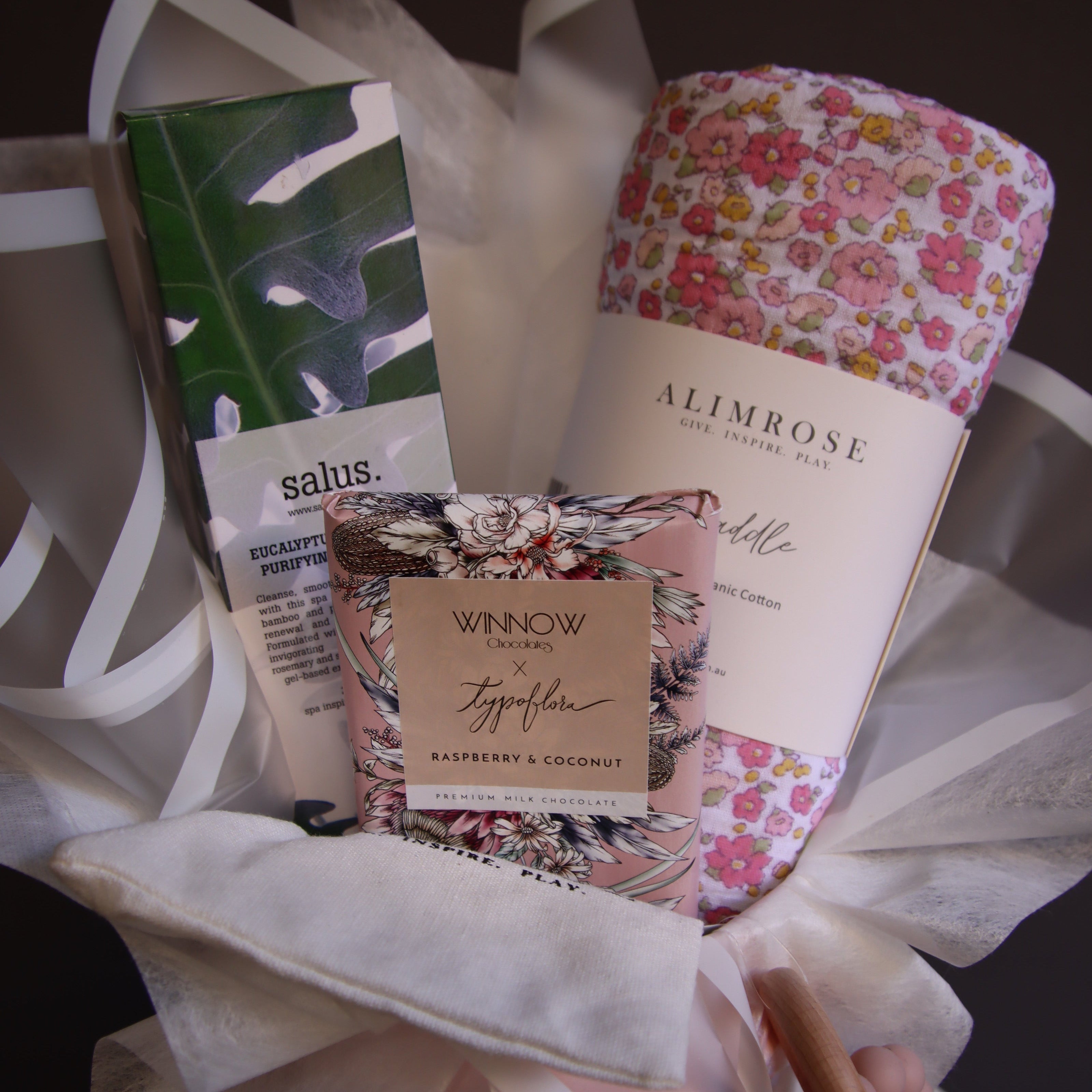 A baby gift set arranged in white tissue paper, featuring a floral-patterned baby blanket, a tube of Salus baby lotion, a Winona brand card, and a pink baby bottle with a wooden spoon.