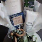 A curated gift basket or bundle containing multiple products including a white tube labeled 'salus', a book or booklet from 'All In Rose', a blue floral patterned item, and a wooden bead teether, arranged in white tissue paper on a dark background.
