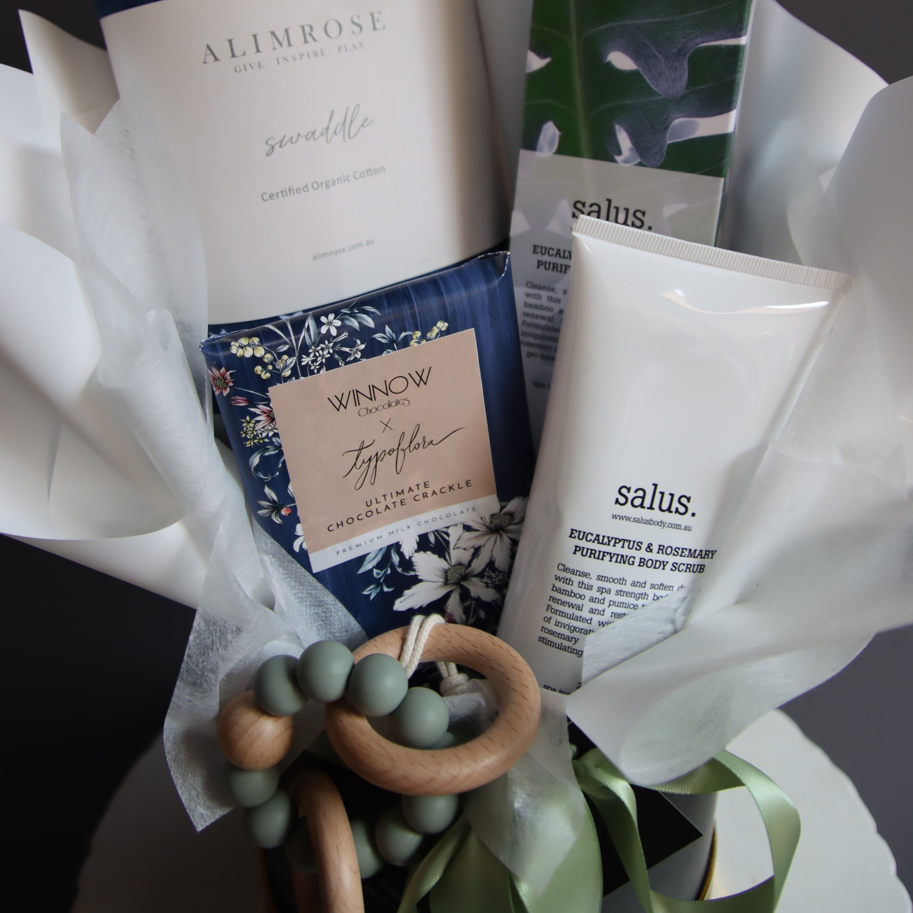 A curated gift basket or bundle containing multiple products including a white tube labeled 'salus', a book or booklet from 'All In Rose', a blue floral patterned item, and a wooden bead teether, arranged in white tissue paper on a dark background.