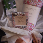 A curated gift set in white tissue paper, featuring a floral-patterned Alimrose roll, a Winkow hand cream tube, a green and white leaf-patterned item, and a peach-colored baby bottle with a wooden spout, tied together with a white ribbon.