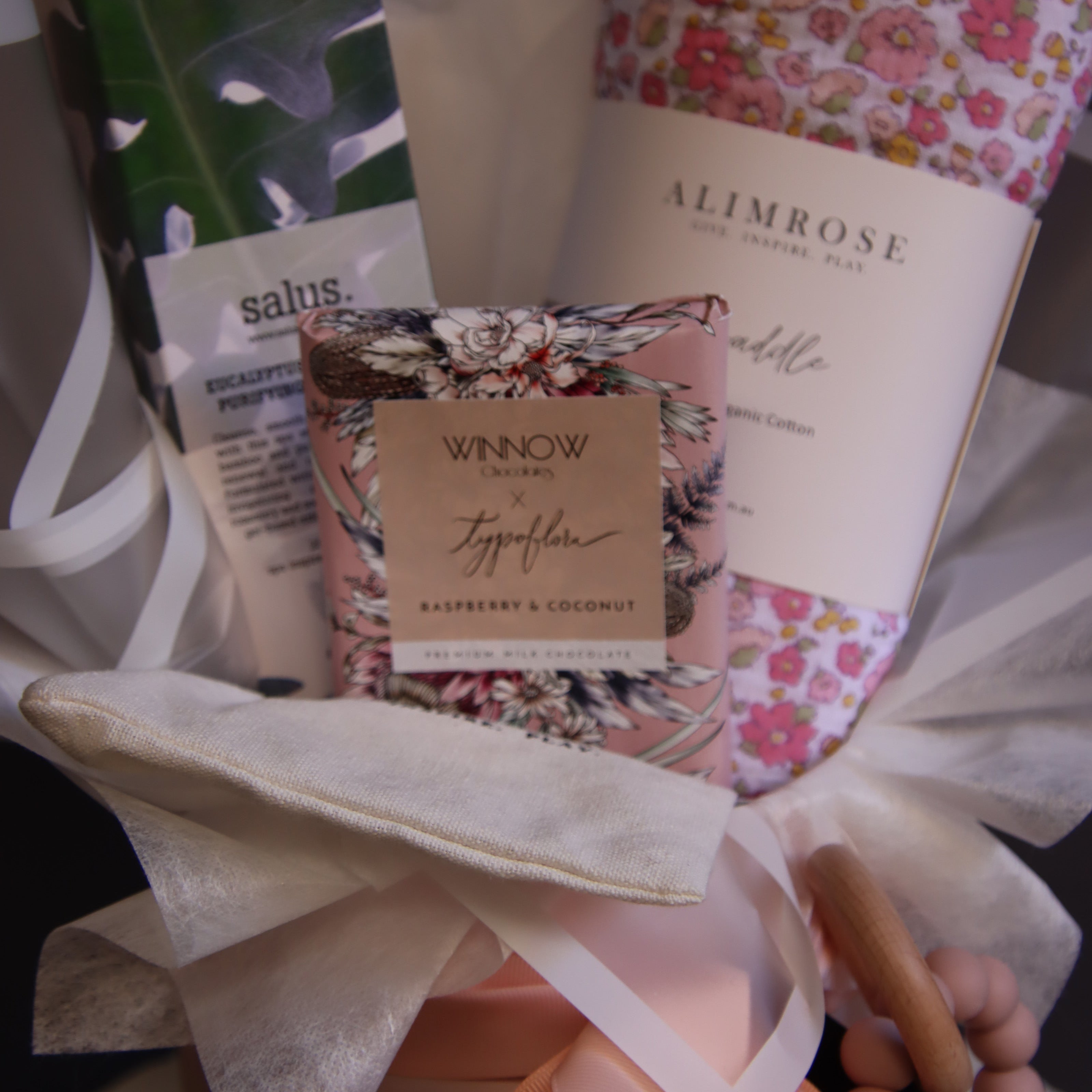 A curated gift set in white tissue paper, featuring a floral-patterned Alimrose roll, a Winkow hand cream tube, a green and white leaf-patterned item, and a peach-colored baby bottle with a wooden spout, tied together with a white ribbon.